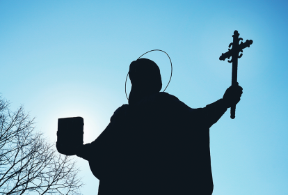 Silhueta da estátua de um santo com os braços abertos - em uma das mãos, ele segura uma cruz; na outra, um livro. O fundo é um céu azul sem nuvens e a silhueta de galhos no canto inferior esquerdo.