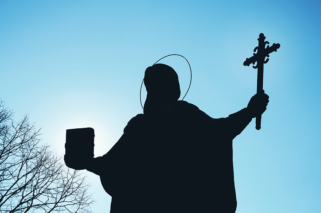 Silhueta da estátua de um santo com os braços abertos - em uma das mãos, ele segura uma cruz; na outra, um livro. O fundo é um céu azul sem nuvens e a silhueta de galhos no canto inferior esquerdo.