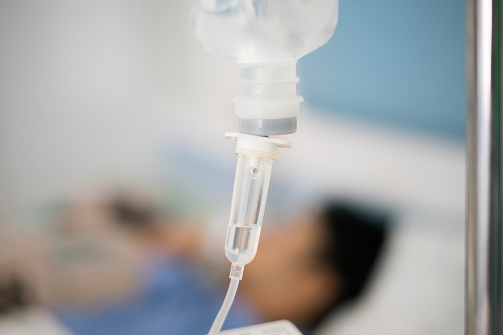 Foco em um dosador de medicamento. Ao fundo, pessoa deitada em uma cama, no hospital.