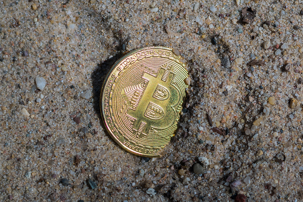 Moeda com símbolo de bitcoin enterrada na areia.