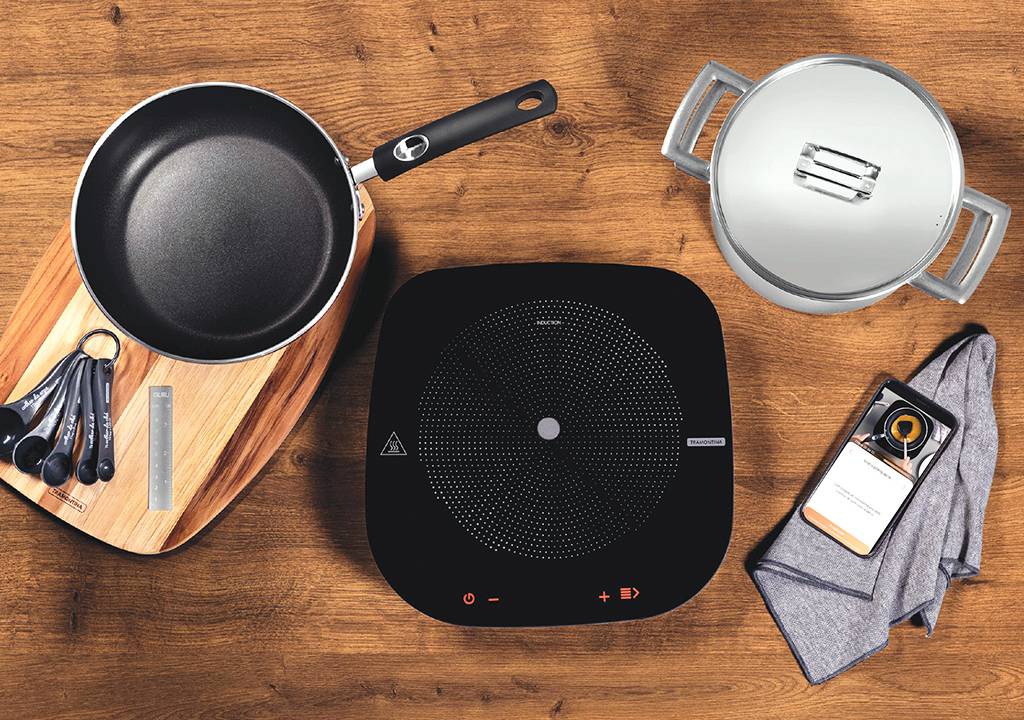 Foto do cooktop Tramontina Guru em cima de uma mesa com duas panelas, um celular, um pano e uma colher de medidas.