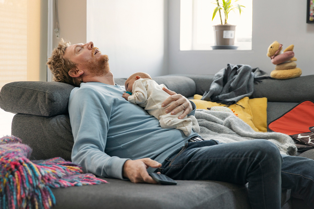 Homem exausto dorme no sofá com bebê em seu colo.