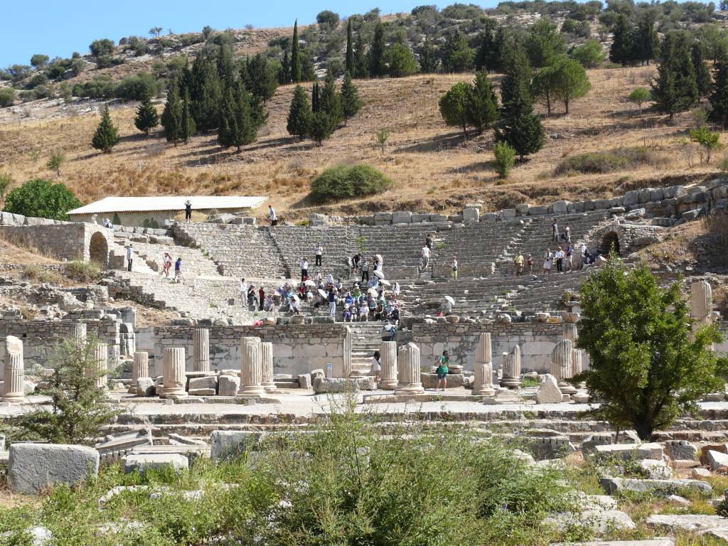 Ephesus_odeon (1) A imagem mostra um antigo anfiteatro de pedra, cinza, sem teto, em ruínas, localizado no sopé de um morro arborizado.