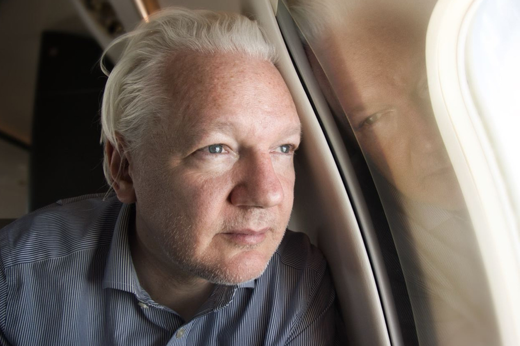 Julian Assange. Homem idoso de cabelo grisalho com o rosto apoiado na janela de um avião.