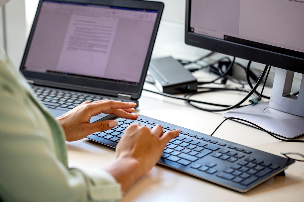Imagem de uma pessoa digitando num teclado, e ao lado um outro computador aberto com um texto já escrito.