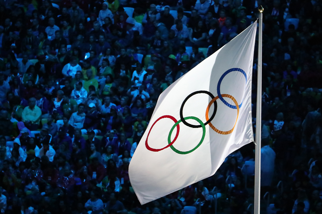 Bandeira com o símbolo olímpico hasteada em frente a uma multidão em uma arquibancada.
