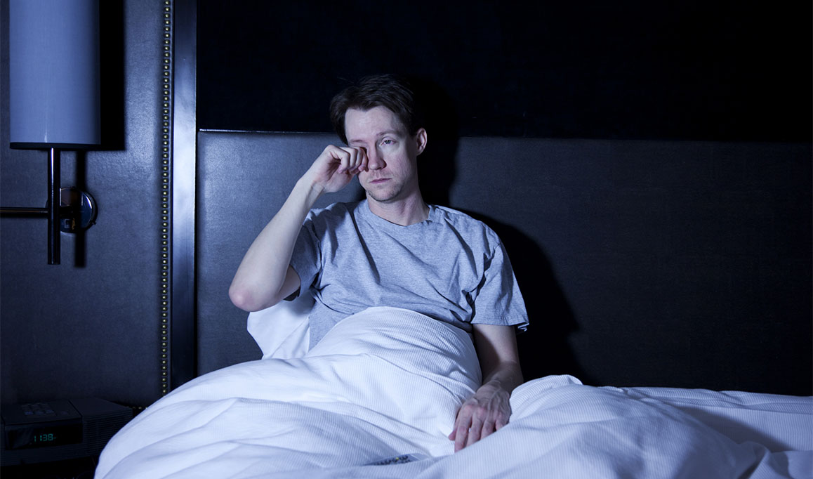 Foto de um homem branco coçando os olhos sentado na cama.