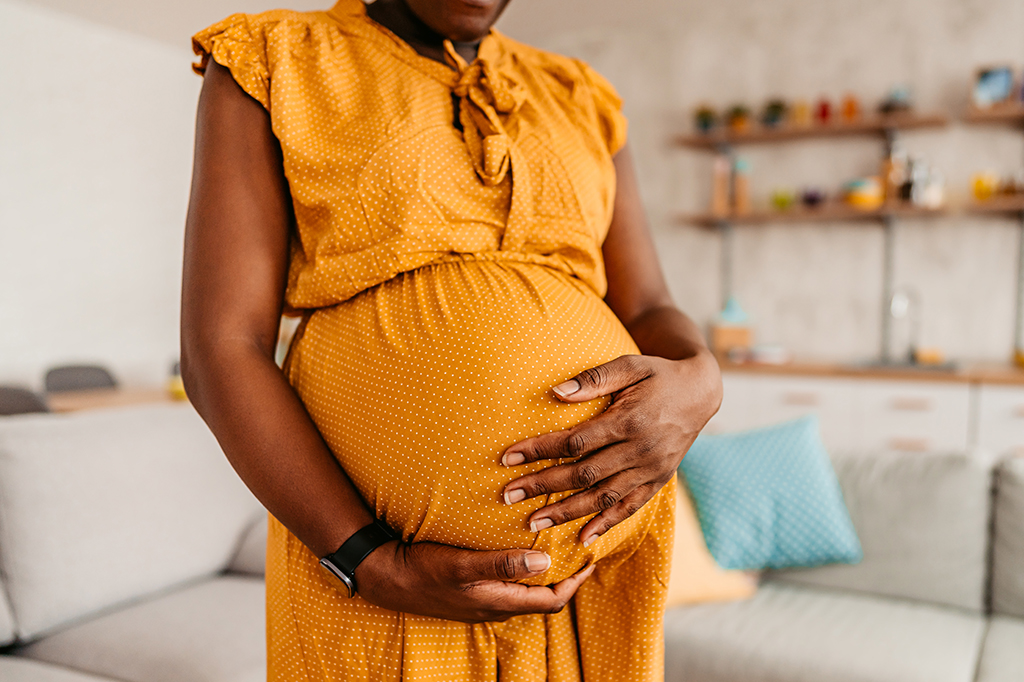 Fotografia de uma mulher negra grávida segurando sua barriga e usando um vestido amarelo em casa.