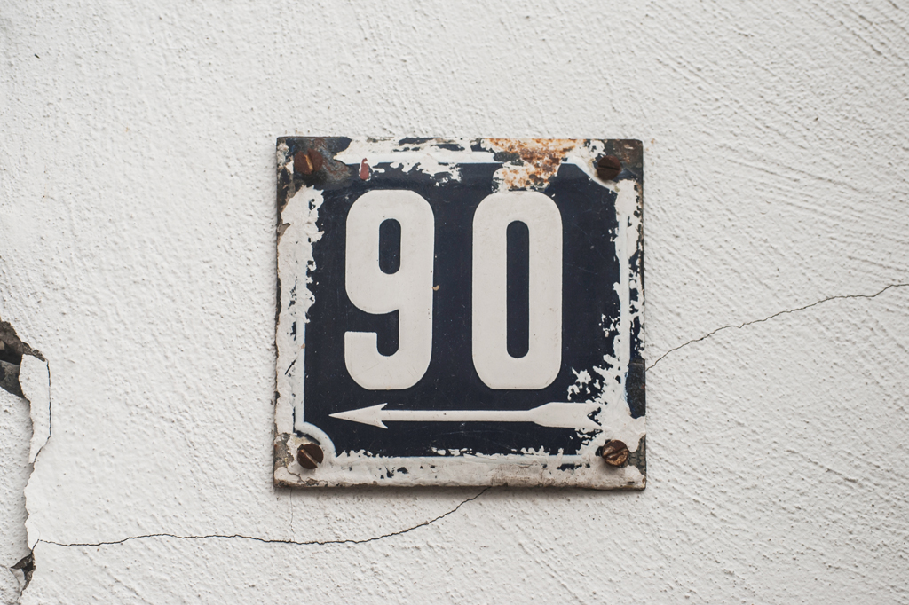 Imagem de uma placa de metal esmaltada quadrada e desgastada, com número de endereço de rua e número 90, parafusada numa rústica parede branca.