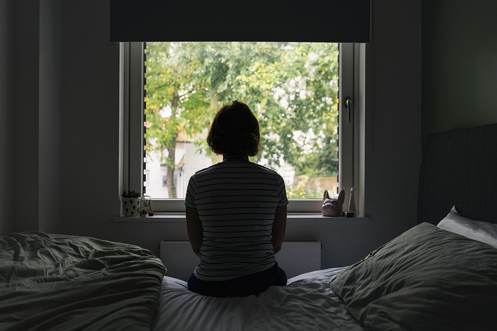Fotografia de costas de uma mulher olhando pela janela do quarto.