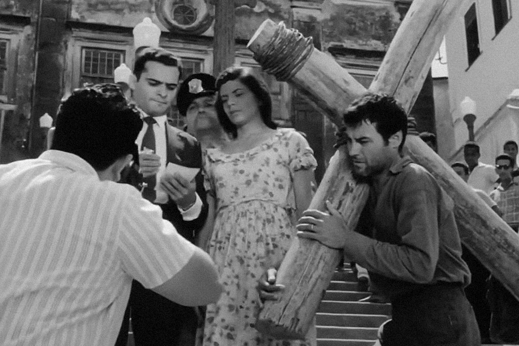 Imagem da cena do filme "O Pagador de Promessas" em que o personagem principal está carregando uma cruz.