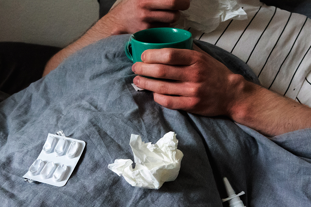 Fotografia de um homem deitado na cama com lenços, xícara de chá, spray nasal e comprimidos.