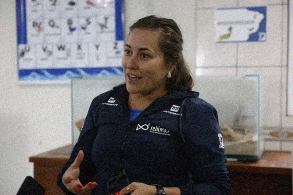 Mulher de cabelos castanhos presos, sorrindo e gesticulando, vestindo jaqueta azul-marinho com logotipos da Petrobras e Rebimar. Ela usa brincos de p&eacute;rola e esmalte vermelho. Ao fundo, um quadro com letras e imagens, e um aqu&aacute;rio sobre um m&oacute;vel de madeira