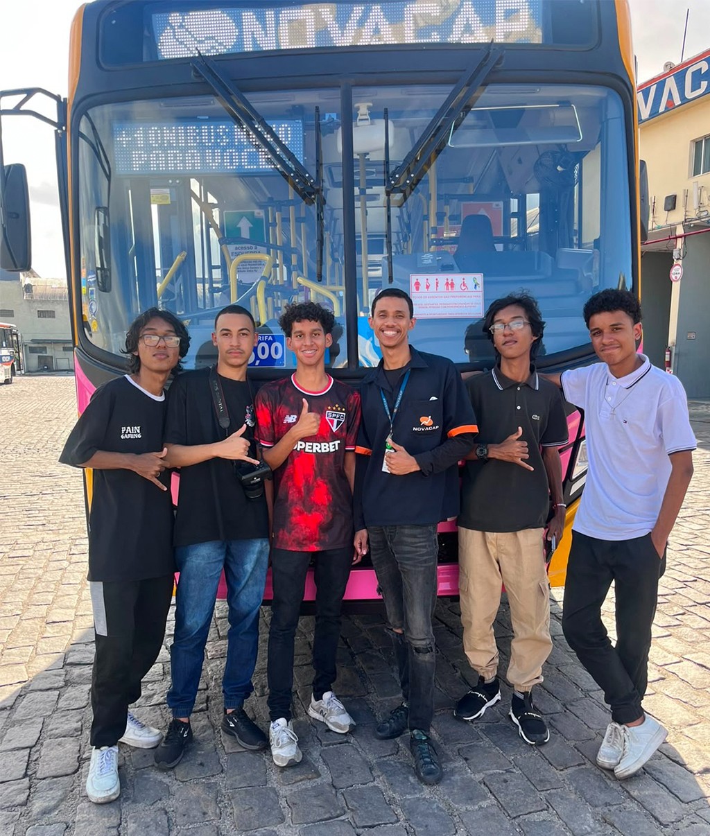 Seis jovens sorridentes, com idades entre 15 e 25 anos, posam em frente a um &ocirc;nibus rosa e amarelo da empresa Novacap. O &ocirc;nibus exibe no letreiro Novacap e Temos um lugar para voc&ecirc;. Os jovens vestem roupas casuais, incluindo camisetas de times de futebol e uma camisa polo. O ch&atilde;o &eacute; de paralelep&iacute;pedos.