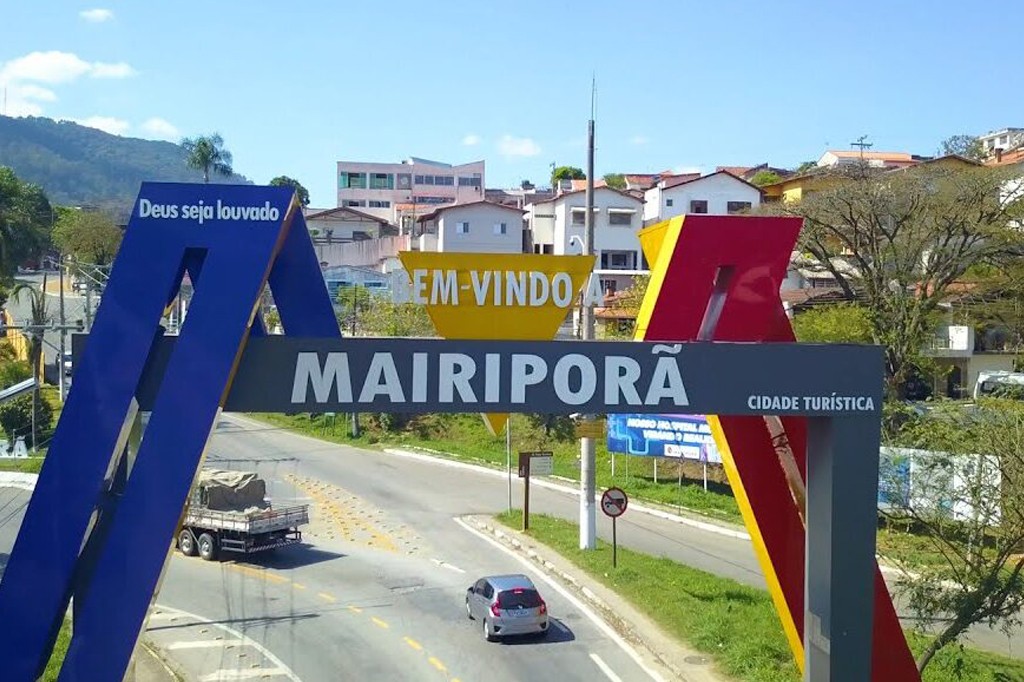 P&oacute;rtico de entrada da cidade de Mairipor&atilde;, com as palavras Deus seja louvado, Bem-vindo a Mairipor&atilde; e Cidade Tur&iacute;stica em destaque. Um caminh&atilde;o e um carro trafegam pela rodovia, com casas e vegeta&ccedil;&atilde;o ao fundo sob c&eacute;u azul.