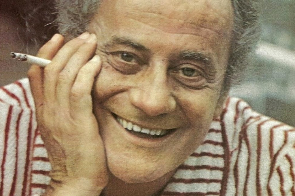Um homem de pele clara, cabelos grisalhos e rosto enrugado, sorri amplamente, com a m&atilde;o direita apoiada na bochecha e um cigarro aceso entre os dedos. Ele veste uma camisa listrada em branco e vermelho. Retrato de Paulo Autran.