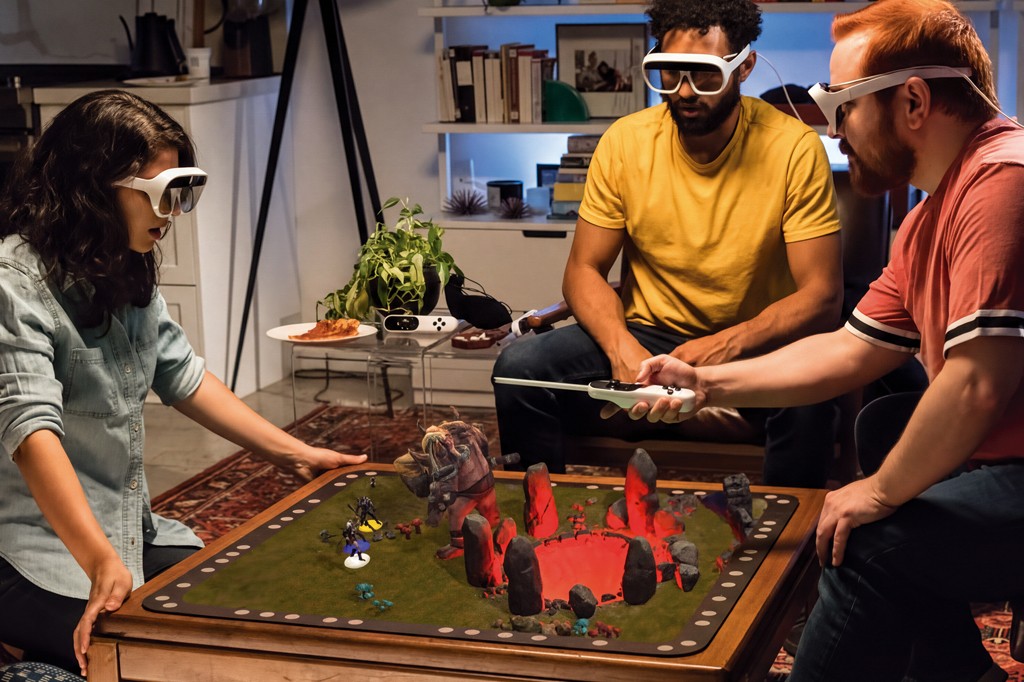 Tr&ecirc;s pessoas, duas sentadas e uma ajoelhada, usam &oacute;culos de realidade aumentada para jogar um jogo de tabuleiro com figuras hologr&aacute;ficas de monstros e guerreiros. O homem ruivo &agrave; direita segura um controle branco, enquanto a mulher &agrave; esquerda e o homem de camiseta amarela observam o tabuleiro com aten&ccedil;&atilde;o. O cen&aacute;rio &eacute; uma sala de estar com estantes e plantas.