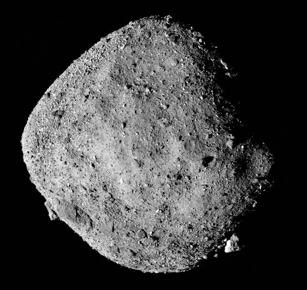 Asteroide Bennu, rochoso e irregular, em tons de cinza, com superf&iacute;cie coberta por pedras e crateras de diversos tamanhos, flutuando no espa&ccedil;o escuro