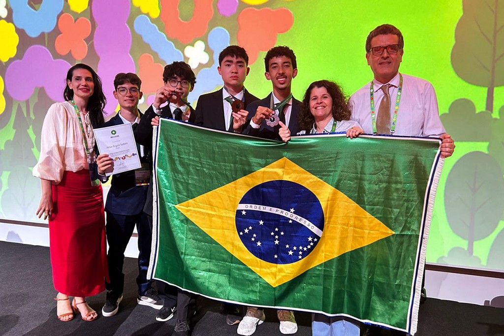 Um grupo de sete pessoas, incluindo quatro jovens e três adultos, sorriem enquanto seguram uma bandeira do Brasil. Os jovens exibem medalhas e um certificado, celebrando uma conquista em um evento com fundo colorido