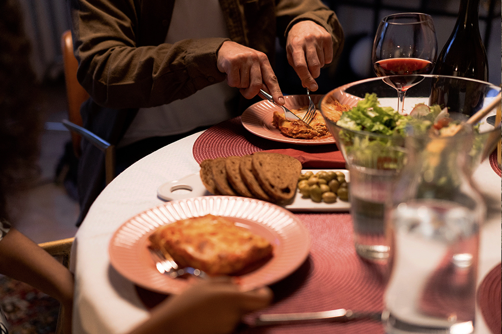 Pessoa de camisa marrom cortando lasanha em prato rosa, com pão, azeitonas, salada e vinho tinto na mesa.