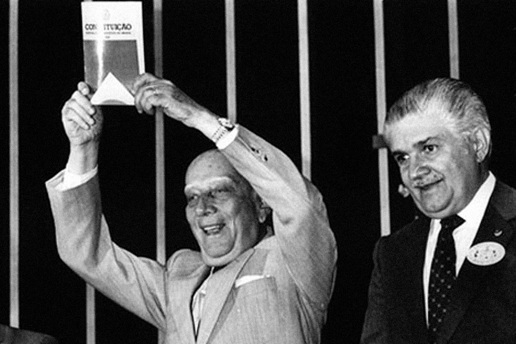 Ulysses Guimarães, careca e sorridente, ergue a Constituição de 1988 com as duas mãos, ao lado de um homem de terno e gravata, também sorrindo, em um fundo escuro com listras verticais claras